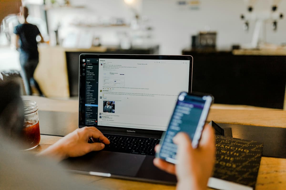Pracownik firmy korzystający jednocześnie z laptopa MacBook Pro z otwartą aplikacją do komunikacji zespołowej i smartfona  – zdalne wsparcie IT i helpdesk dla firm w działaniu.   